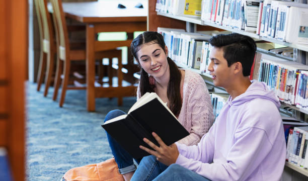 Two 16 year old high school students in the library. A multiracial teenage boy, Asian and Caucasian, is sitting on the floor, holding up a book to show to his friend, a teenage girl next to him.