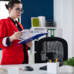 businesswoman-standing-up-desk-holding-clipboard-with-chart-focused-entrepreneur-with-glasses-red-jacket-startup-office-looking-business-results-computer-screen