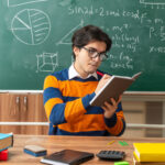 concentrated-young-geometry-teacher-wearing-glasses-sitting-desk-with-school-supplies-classroom-writing-note-pad-with-pen
